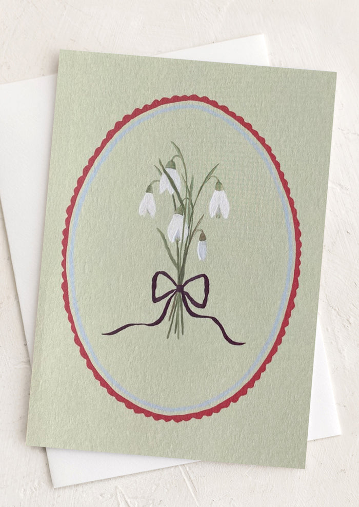 A sage green greeting card with oval frame around snowdrop flowers.