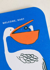 A blue greeting card with illustration of pelican with baby pelican in its beak, text reads "Welcome, baby".